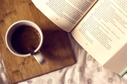 [ai] A white coffee mug filled with dark coffee placed on a wooden board next to an open book on a patterned tablecloth. The pages of the book are visible.
